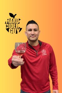 Adrian Luna holding a glass of cider with The Hard Cider Guy logo visible