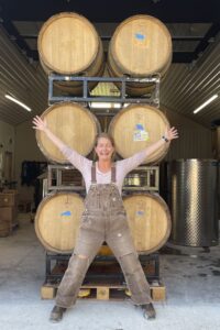 Martha Lowry of Woodfolk Cider standing with arms outstretched in front of stacked oak barrels at the cidery in Ontario, Canada.