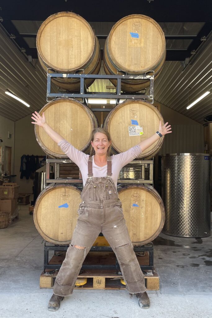 Martha Lowry of Woodfolk Cider standing with arms outstretched in front of stacked oak barrels at the cidery in Ontario, Canada.