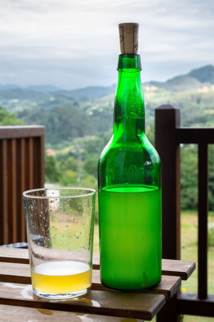 A bottle of traditional Asturian cider beside a glass overlooking the hills of Asturias, Spain.